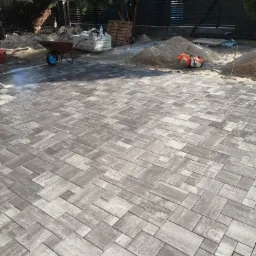 Ułożona, szara kostka brukowa o nieregularnym wzorze, widoczna taczka z niebieskim kołem, paleta z kostką, piasek i przecinarka do kostki w tle.