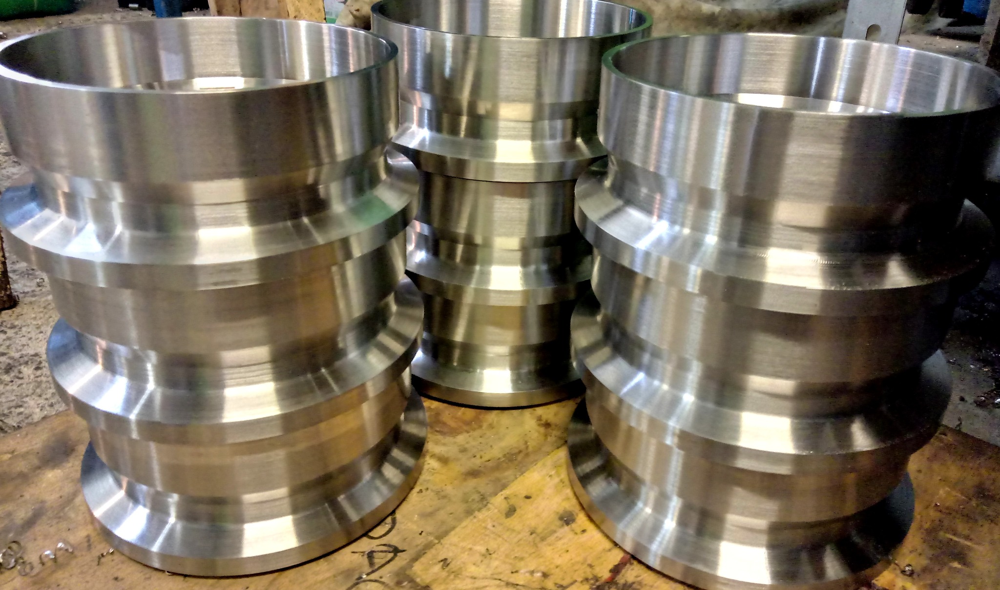 Trzy metalowe elementy o kształcie cylindrycznym z rowkami, ułożone na drewnianej powierzchni. Widoczne ślady obróbki i refleksy światła na metalu.