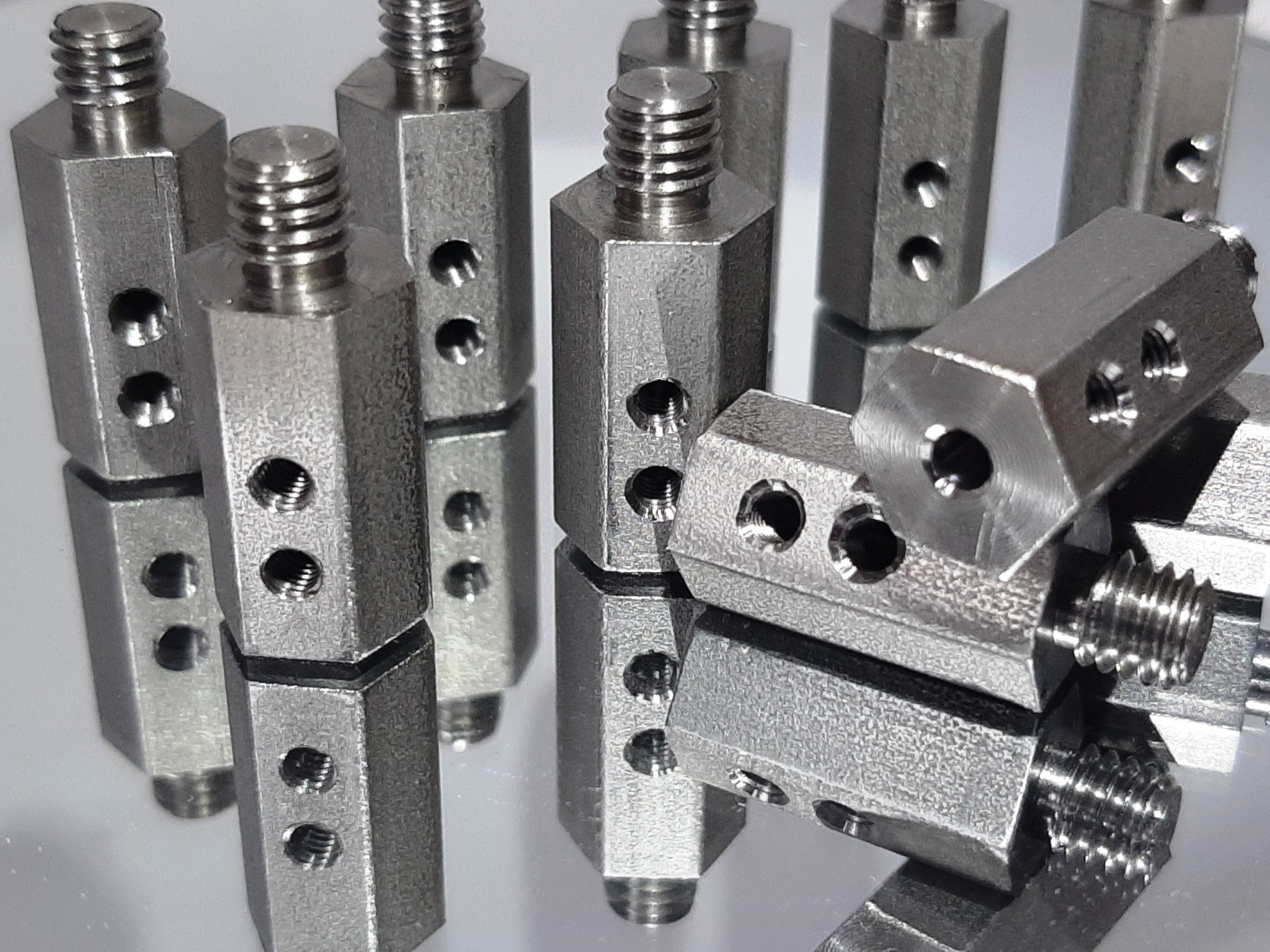 Seria metalowych, sześciokątnych elementów z gwintem i otworami, ułożonych na odblaskowej powierzchni, ukazujących precyzję wykonania.