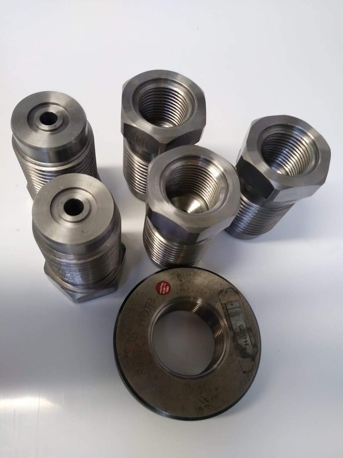 Sześć metalowych nakrętek z gwintem wewnętrznym, niektóre z sześciokątną podstawą, ułożonych na białym tle. Na jednej z nakrętek widoczny czerwony znak i numer seryjny.