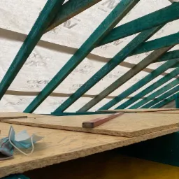 Konstrukcja dachu z pomalowanymi na zielono belkami, widoczna izolacja oraz płyty OSB, poziomica, rękawice robocze i maseczka leżą na płytach.