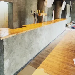 Surowy bar z drewnianym blatem i betonowym frontem w trakcie prac wykończeniowych, widoczne dystrybutory do piwa i surowe ściany w tle, podłoga częściowo zabezpieczona folią.