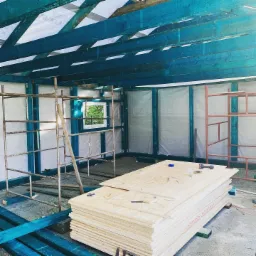 Wnętrze w trakcie budowy: szkielet domu z pomalowanymi na niebiesko elementami konstrukcyjnymi, rusztowania i stos płyt OSB z narzędziami na wierzchu, widok na okno z zielenią na zewnątrz.