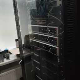 Serwerownia: szafa rack z zainstalowanymi urządzeniami sieciowymi, widoczne kable, switche, routery i panele krosownicze w szklanej obudowie, obok okno.