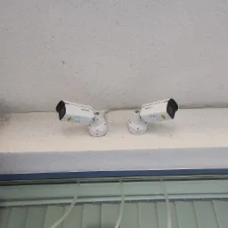 Dwie białe kamery monitoringu Hikvision zamontowane na suficie, widoczne okno z żaluzjami w tle.