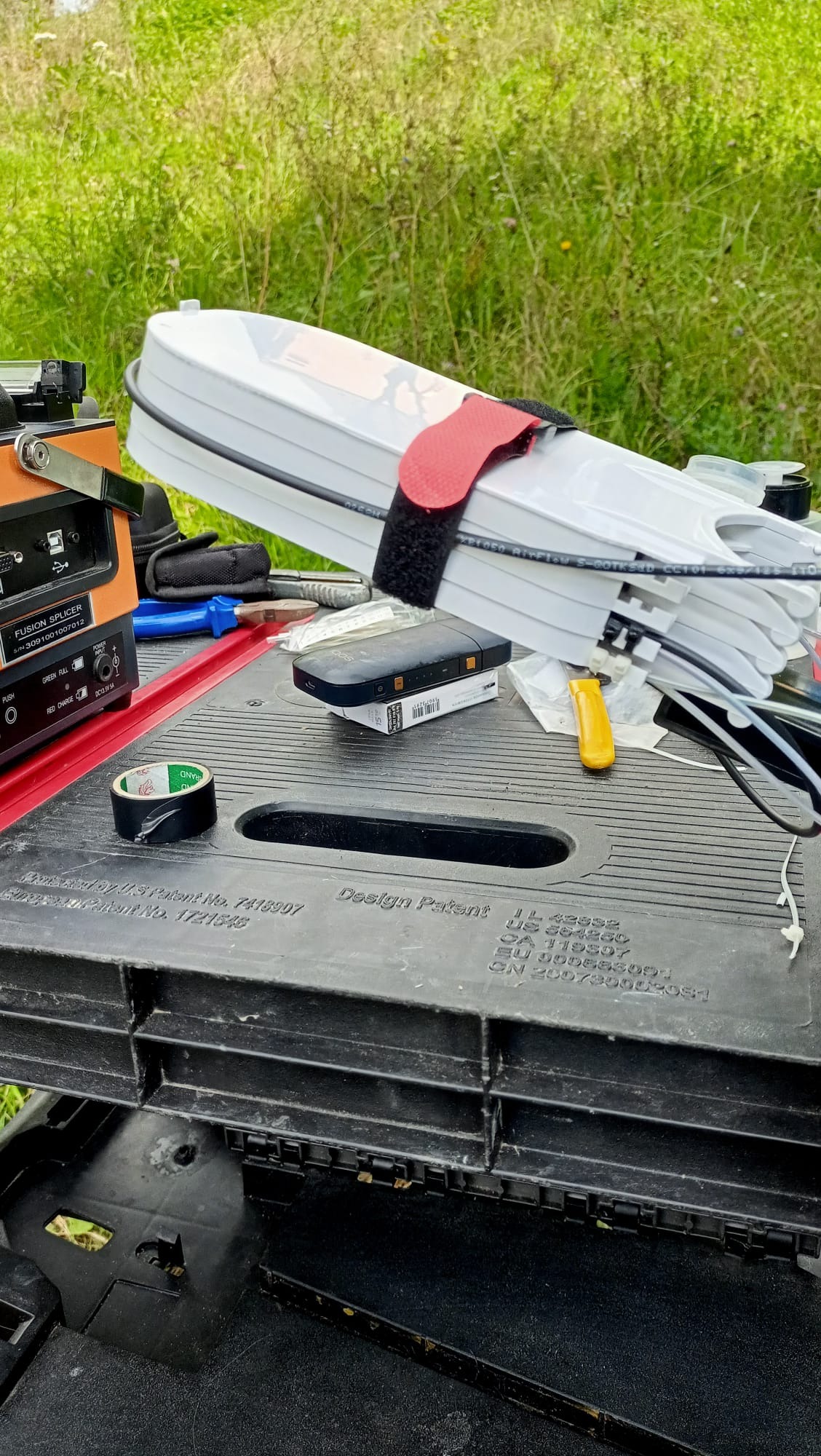 Przygotowane kable światłowodowe w białej osłonie z czerwonym paskiem na czarnym stole roboczym z narzędziami, zgrzewarką światłowodową oraz zieloną trawą w tle.