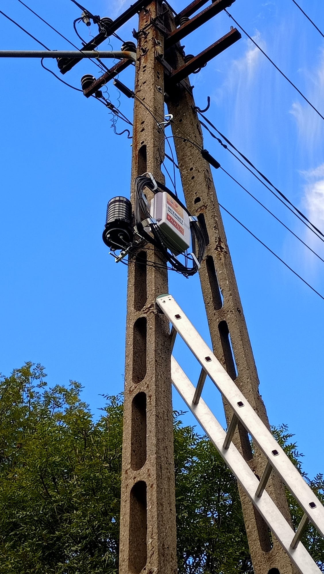 Betonowy słup telekomunikacyjny z zamontowanym sprzętem sieciowym i przystawioną aluminiową drabiną na tle błękitnego nieba.