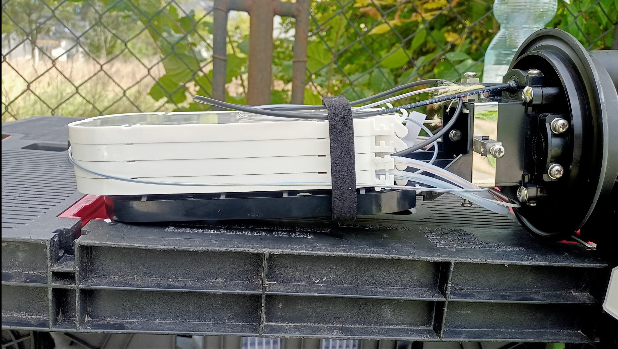 Złącze światłowodowe z widocznymi kablami i elementami montażowymi, umieszczone na czarnym, plastikowym stole roboczym na zewnątrz.