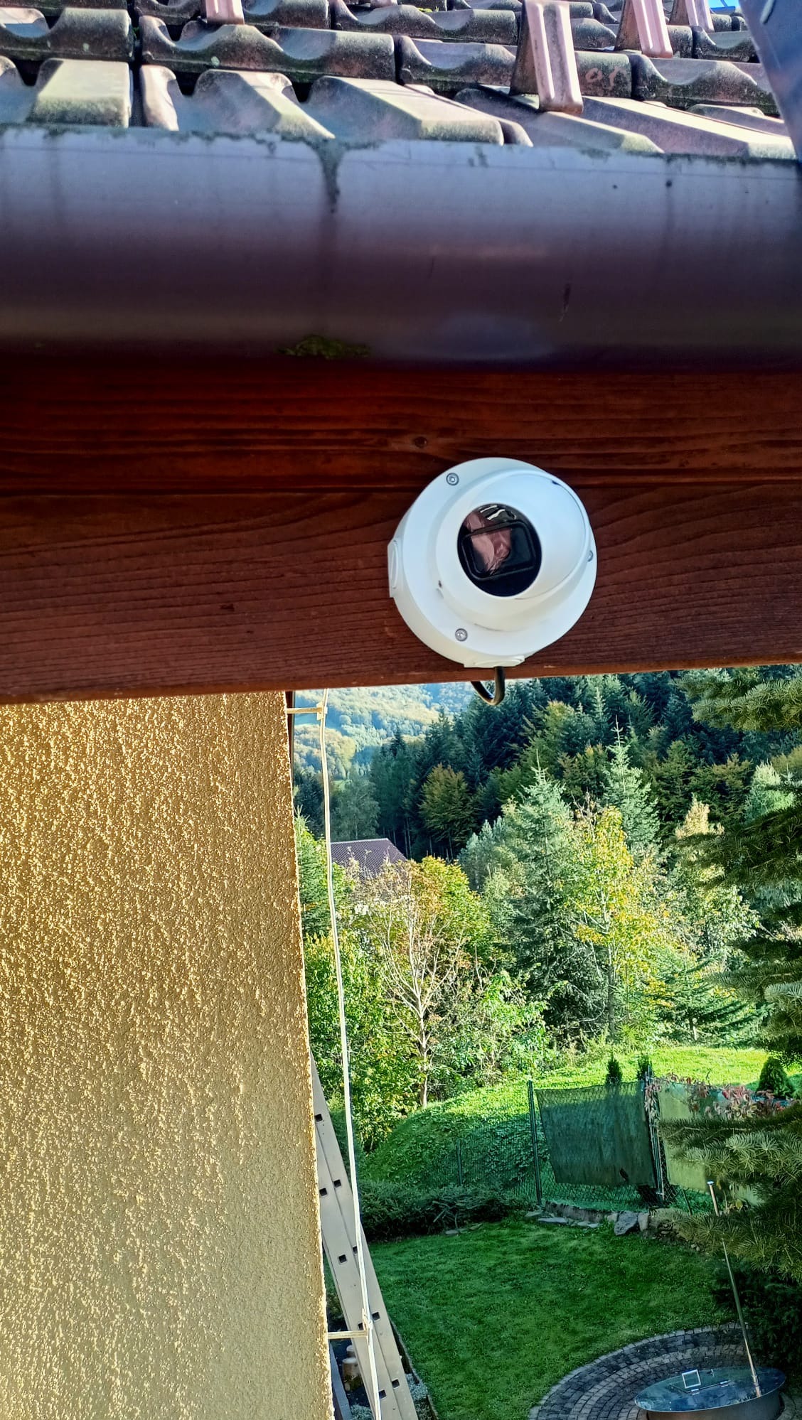 Kamera monitoringu zamontowana pod okapem domu z widokiem na ogród i drzewa w tle, drabina oparta o ścianę budynku.