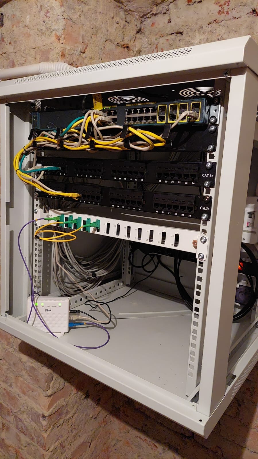 Szafa rack z zamontowanym switchem, patch panelem Cat.5e oraz routerem ZTE z podłączonymi kablami Ethernet i światłowodowymi na tle ceglanej ściany w piwnicy.