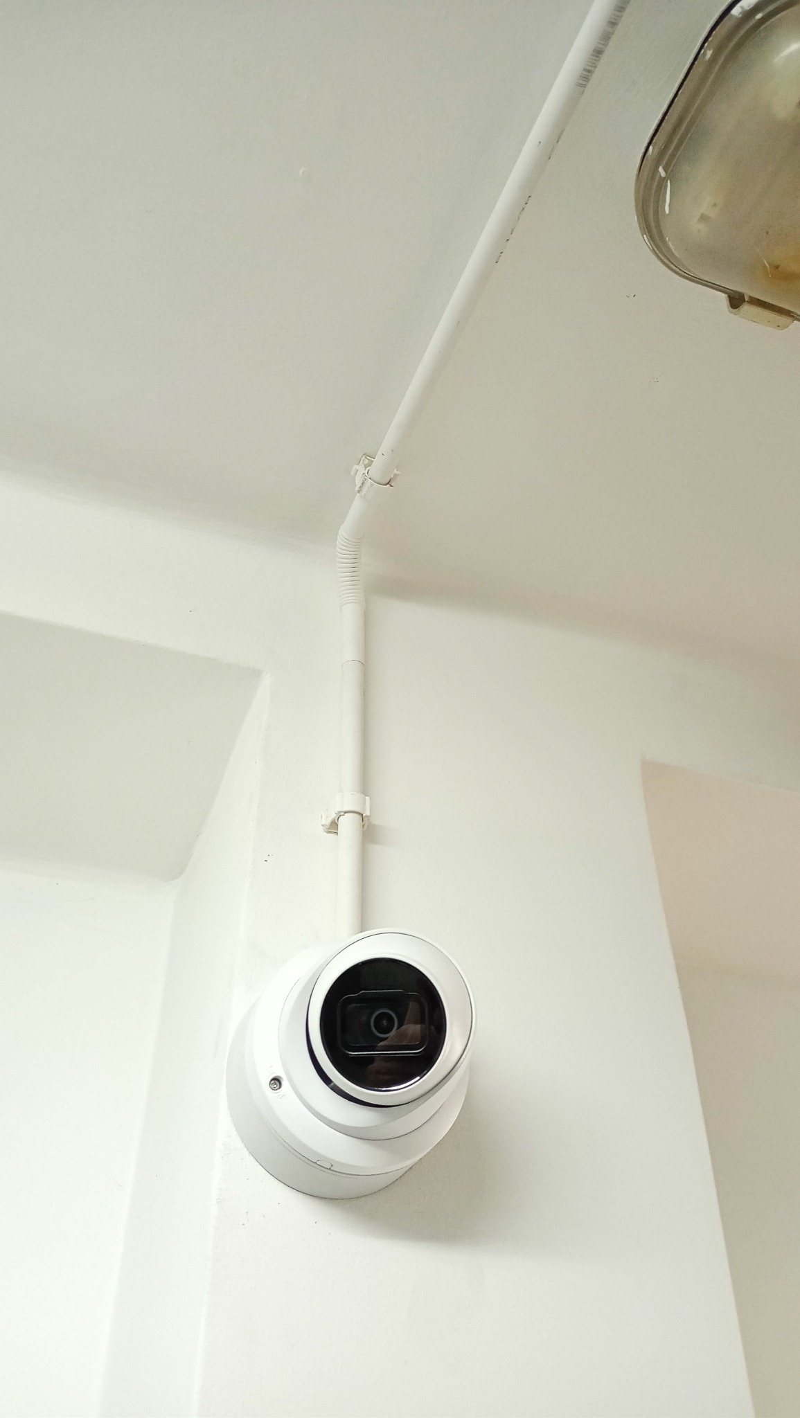 Kopułkowa kamera monitoringu CCTV zamontowana na białej ścianie, z widoczną białą rurką instalacyjną prowadzącą do sufitu i lampą w tle.