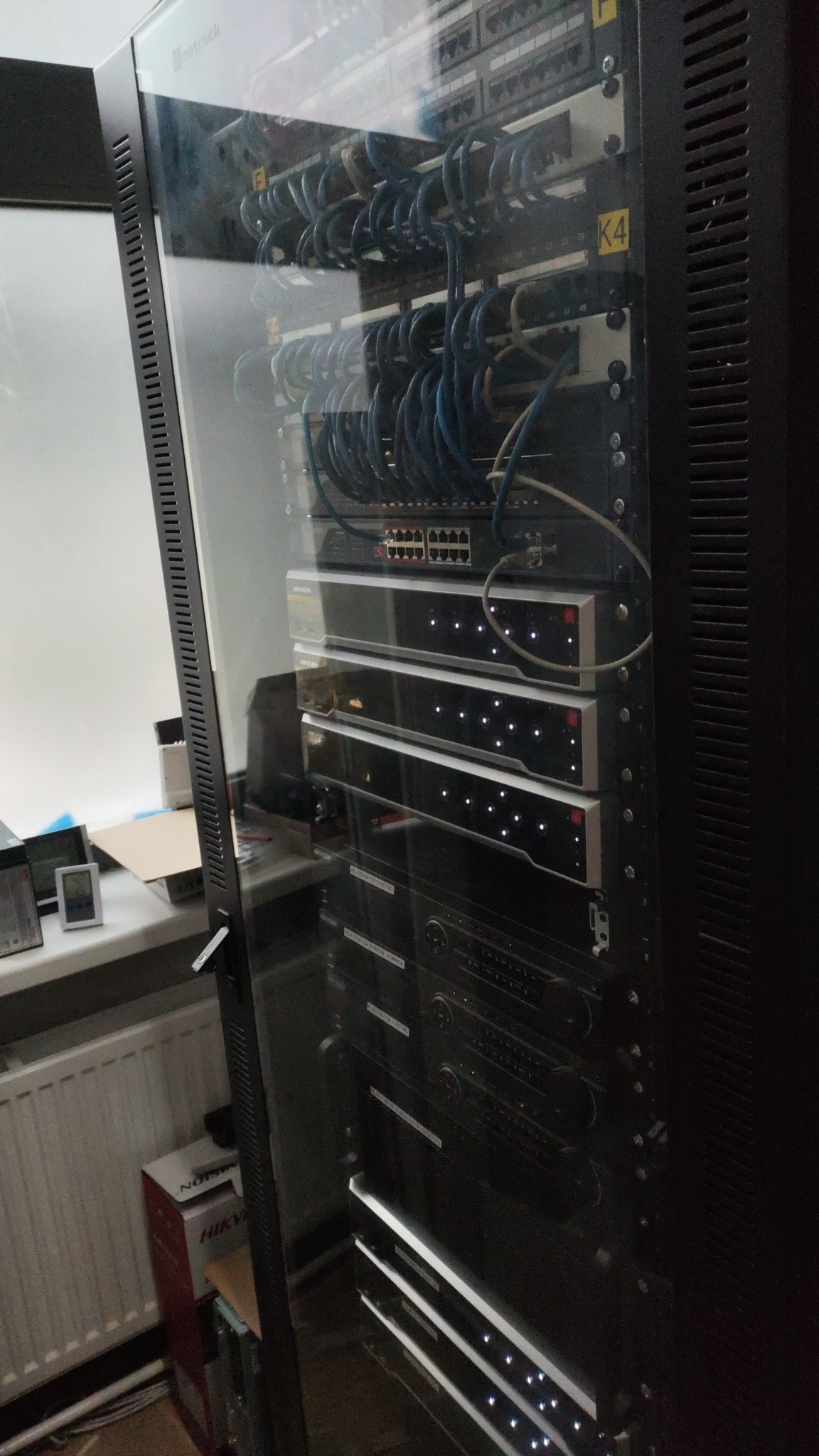 Serwerownia: szafa rack z zainstalowanymi urządzeniami sieciowymi, widoczne kable, switche, routery i panele krosownicze w szklanej obudowie, obok okno.