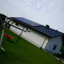 Dom jednorodzinny z panelami fotowoltaicznymi na dachu, widok z boku z perspektywy trawnika, na pierwszym planie huśtawka typu bocianie gniazdo.