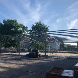 Wznoszenie konstrukcji stalowej hali na betonowym fundamencie, widoczne elementy rusztowania, w tle budynek z białej cegły i drzewa pod błękitnym niebem.