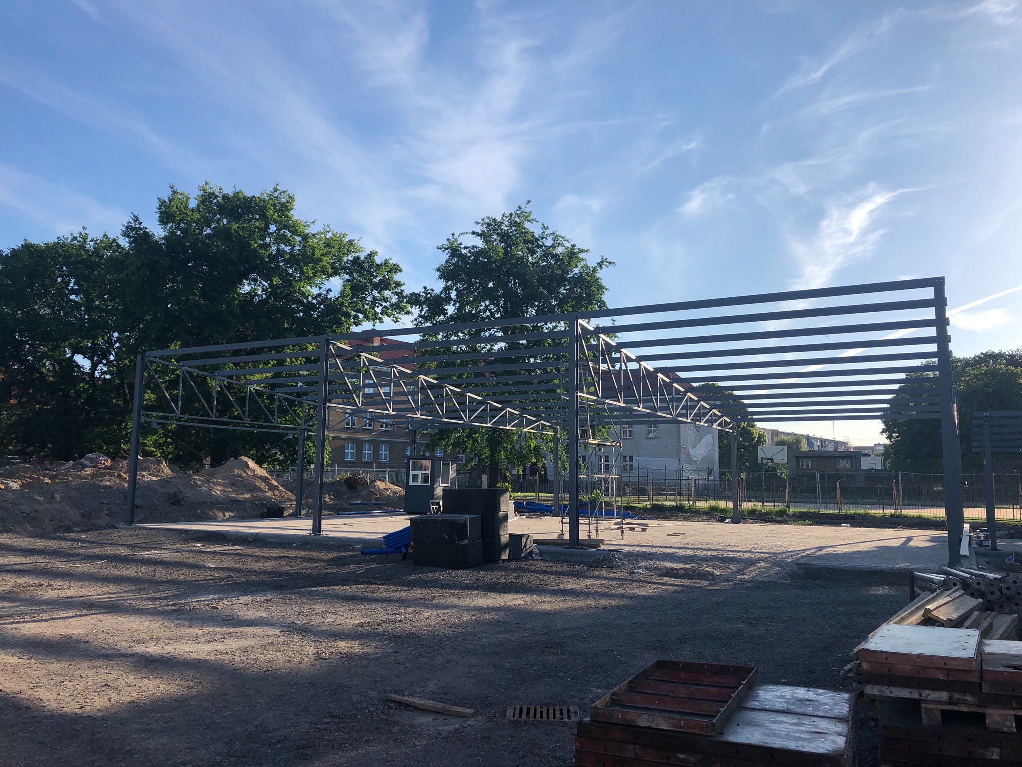 Wznoszenie konstrukcji stalowej hali na betonowym fundamencie, widoczne elementy rusztowania, w tle budynek z białej cegły i drzewa pod błękitnym niebem.