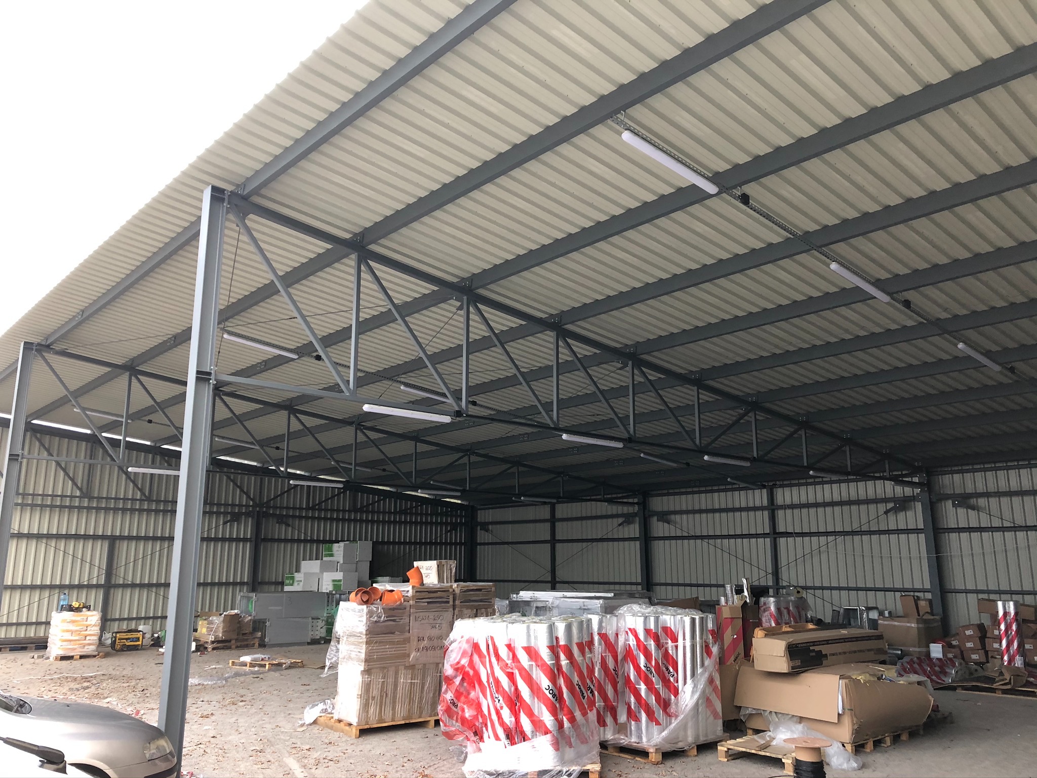 Wnętrze hali w trakcie budowy z widoczną stalową konstrukcją, dachem z blachy trapezowej i materiałami budowlanymi składowanymi wewnątrz, częściowo widoczny samochód przed halą.