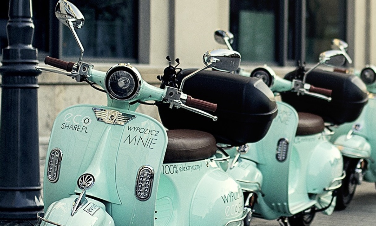 Szereg miętowych skuterów elektrycznych z napisem 'Wypożycz Mnie' i logo 'Eco Share.pl' zaparkowanych przy ulicznej latarni.