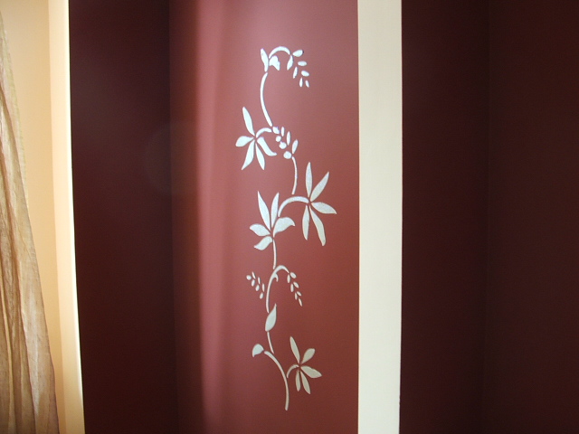 Ciemnoczerwona ściana z ręcznie malowanym, srebrnym motywem roślinnym w stylu secesyjnym, widoczny fragment zasłony po lewej stronie.