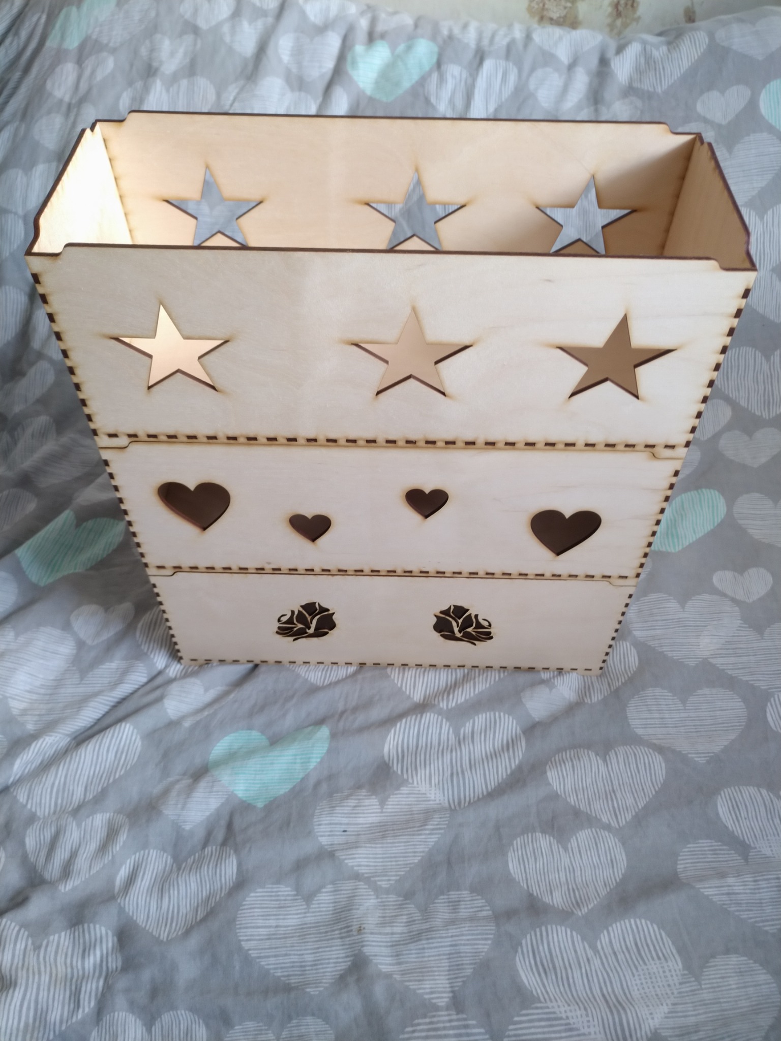 Ozdobny, drewniany koszyk z wyciętymi wzorami gwiazd, serc i róż, ustawiony na pościeli w szare serca.