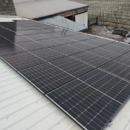 Rząd czarnych paneli słonecznych na metalowym dachu z widoczną białą furgonetką i drzewami w tle.