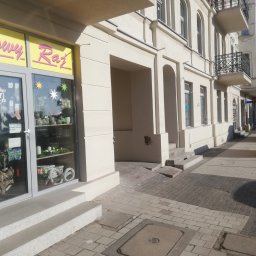 Firma ogólnobudowlana - Fasada kamienicy po renowacji z widocznym odnowionym wejściem do sklepu 'Cenowy Raj', żółty kontener budowlany SKIP na chodniku w tle.