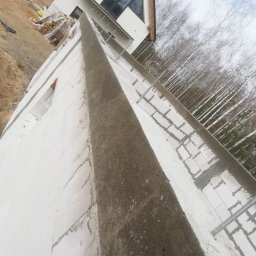 Firma ogólnobudowlana - Wznoszenie muru z bloczków betonu komórkowego Ytong Silka z widocznym zbrojeniem i zalewanym betonem, w tle nowy dom z dużym trójkątnym oknem i las brzozowy.