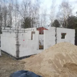 Wznoszenie ścian parterowego domu z białych bloczków betonu komórkowego, widoczne zbrojenia pionowe, sterta piasku na pierwszym planie, drzewa w tle.