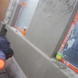 Świeżo nałożona szpachla na ścianie zewnętrznej budynku, widoczne zakryte folią okna z pomarańczową ramą, butelki z napojami stojące na parapecie, fragment ubrania osoby pracującej.