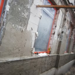 Firma ogólnobudowlana - Elewacja budynku w trakcie prac: nałożona warstwa szpachli z siatką, okna zabezpieczone folią, widoczna puszka z pianką montażową i narzędzia na parapecie.