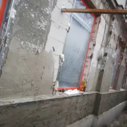 Ocieplanie elewacji budynku w trakcie prac: widoczne okna zabezpieczone folią, siatka pod tynk, świeżo nałożona warstwa zaprawy murarskiej, pianka montażowa uszczelniająca, narzędzia i materiały...