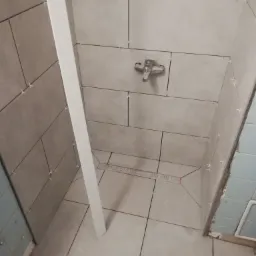 Płytki ceramiczne w trakcie układania w kabinie prysznicowej, widoczne krzyżyki dystansowe, odpływ liniowy i bateria, fragment ściany z niebieskimi płytkami w tle.