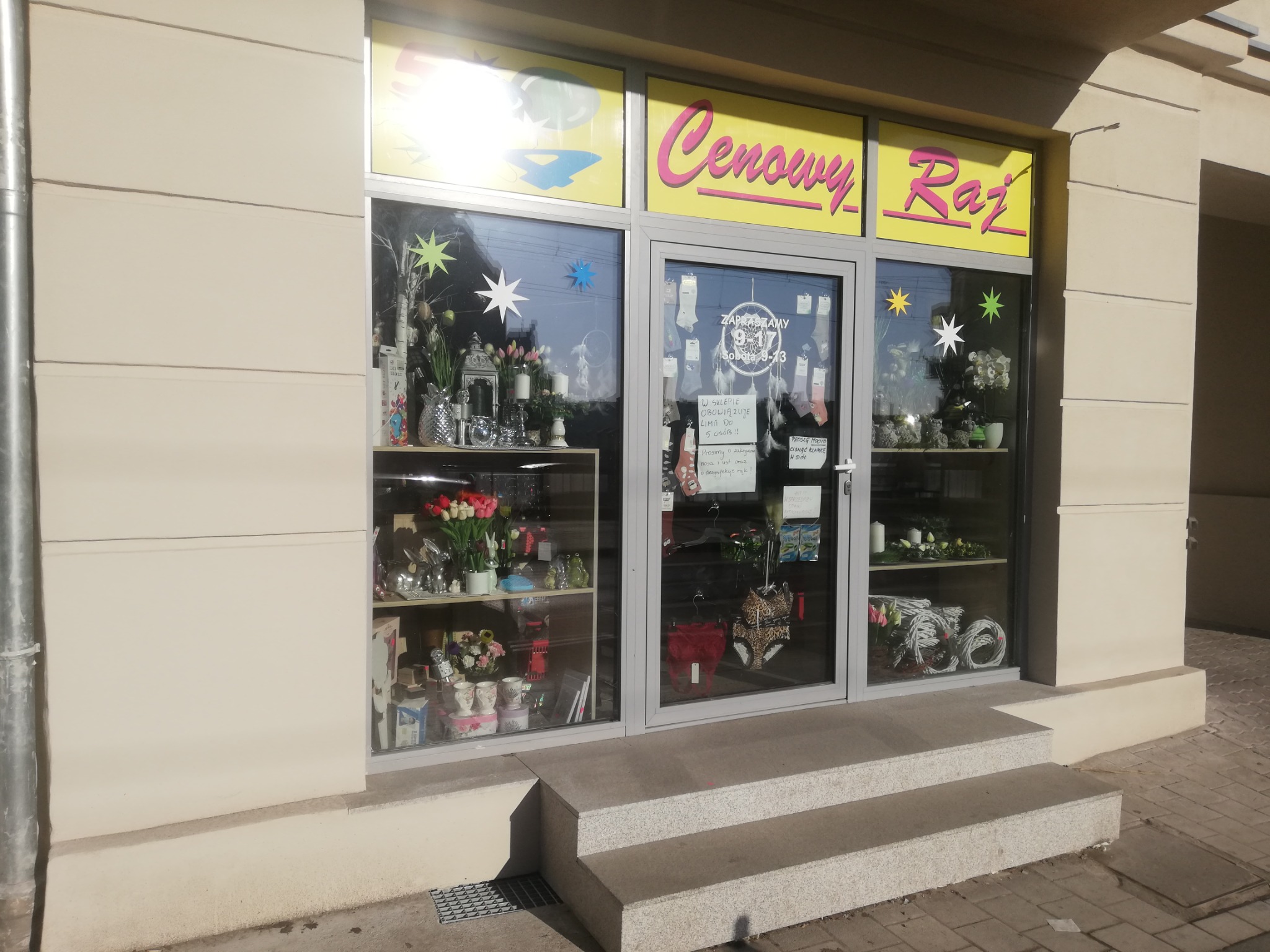 Fasada sklepu z dużymi witrynami, na których widoczne są dekoracje i towary, nad witrynami żółty szyld z napisem 'Cenowy Raj', wejście po schodach.