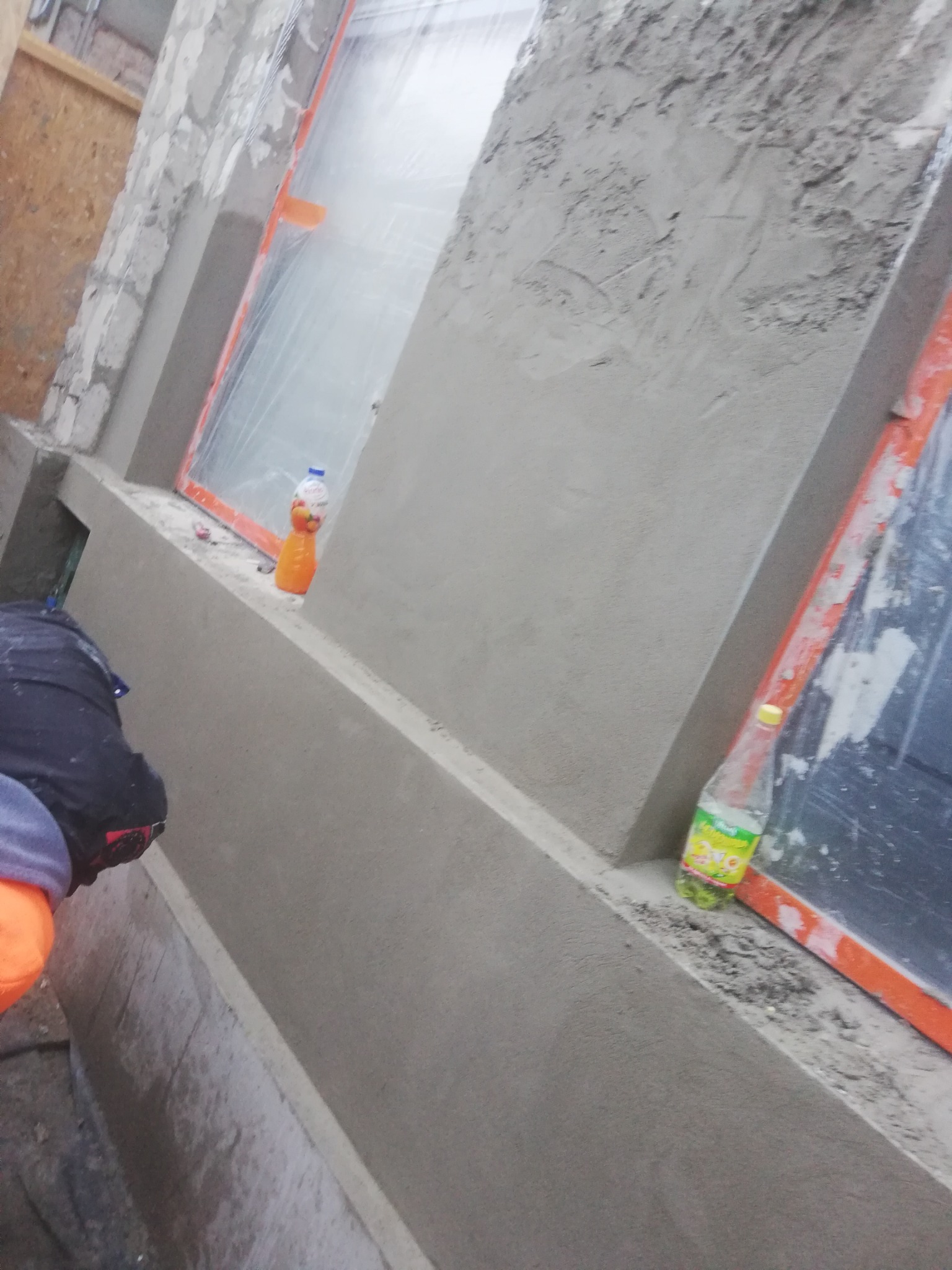 Świeżo nałożona szpachla na ścianie zewnętrznej budynku, widoczne zakryte folią okna z pomarańczową ramą, butelki z napojami stojące na parapecie, fragment ubrania osoby pracującej.