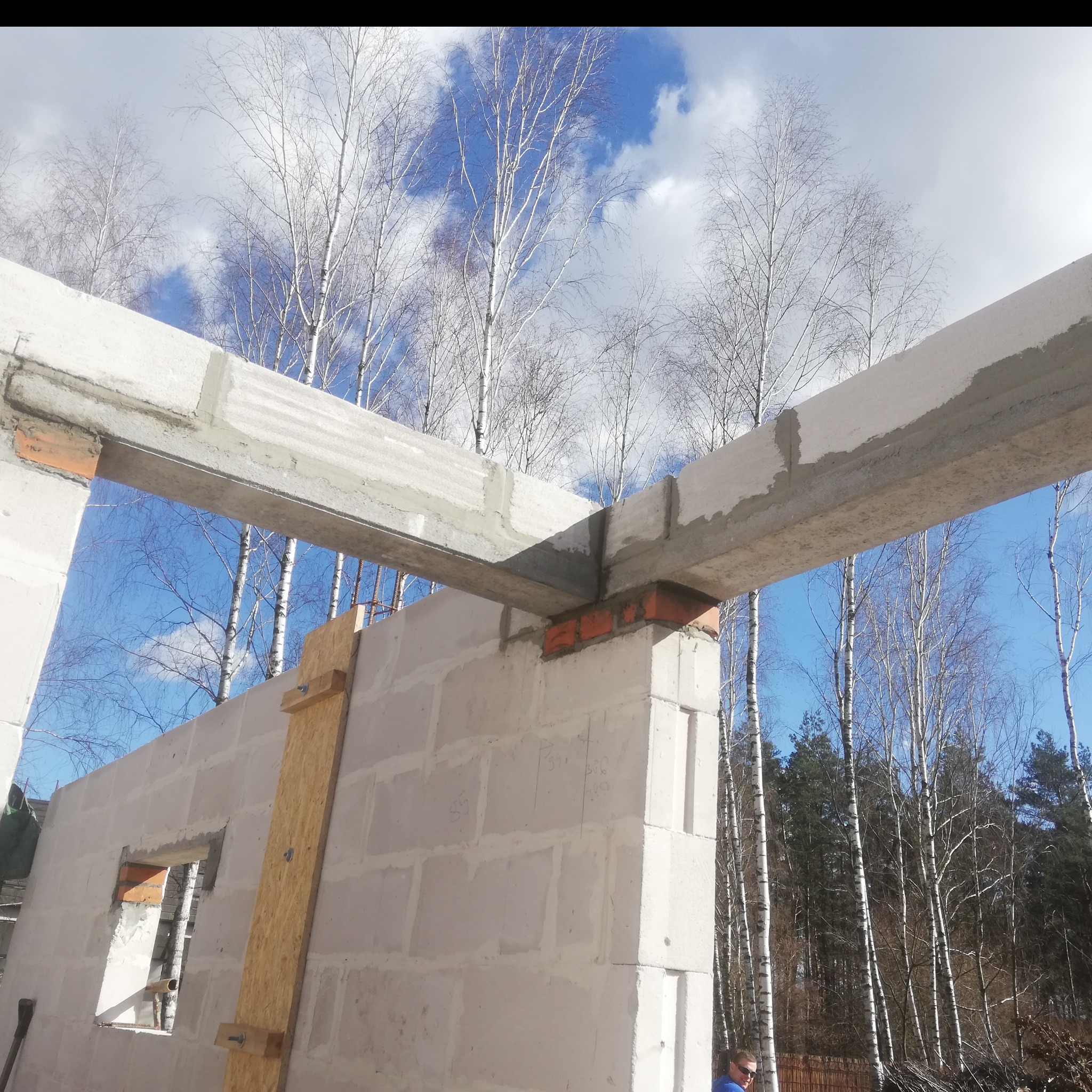 Wznoszenie murów z betonu komórkowego Ytong z nadprożami zbrojonymi, widoczne drewniane stemple podtrzymujące nadproże, w tle las brzozowy i błękitne niebo.