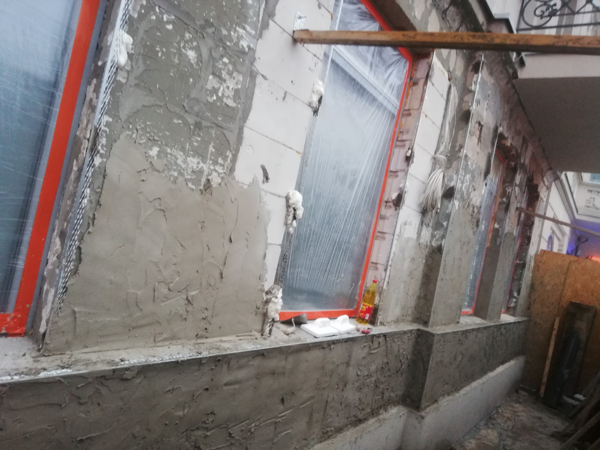 Ocieplanie elewacji budynku w trakcie prac: widoczne okna zabezpieczone folią, siatka pod tynk, świeżo nałożona warstwa zaprawy murarskiej, pianka montażowa uszczelniająca, narzędzia i materiały...