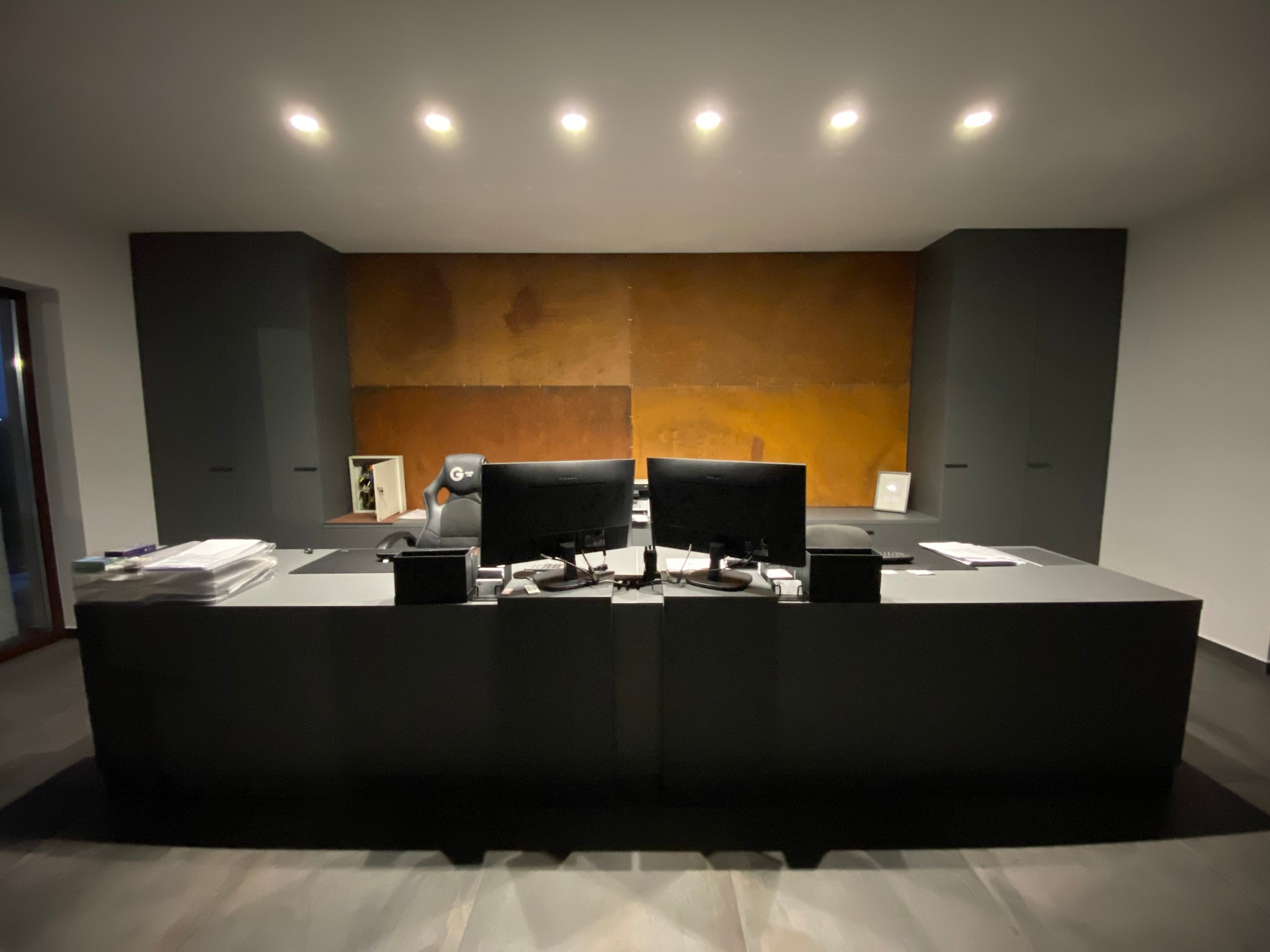 Nowoczesne biuro z dużym, ciemnym biurkiem, dwoma monitorami, brązową ścianą w tle i dyskretnym oświetleniem punktowym.