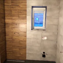 Ściana łazienki wyłożona płytkami imitującymi drewno sąsiaduje z szarymi płytkami, widoczne otwory na armaturę i okno z widokiem na niebieski dach.