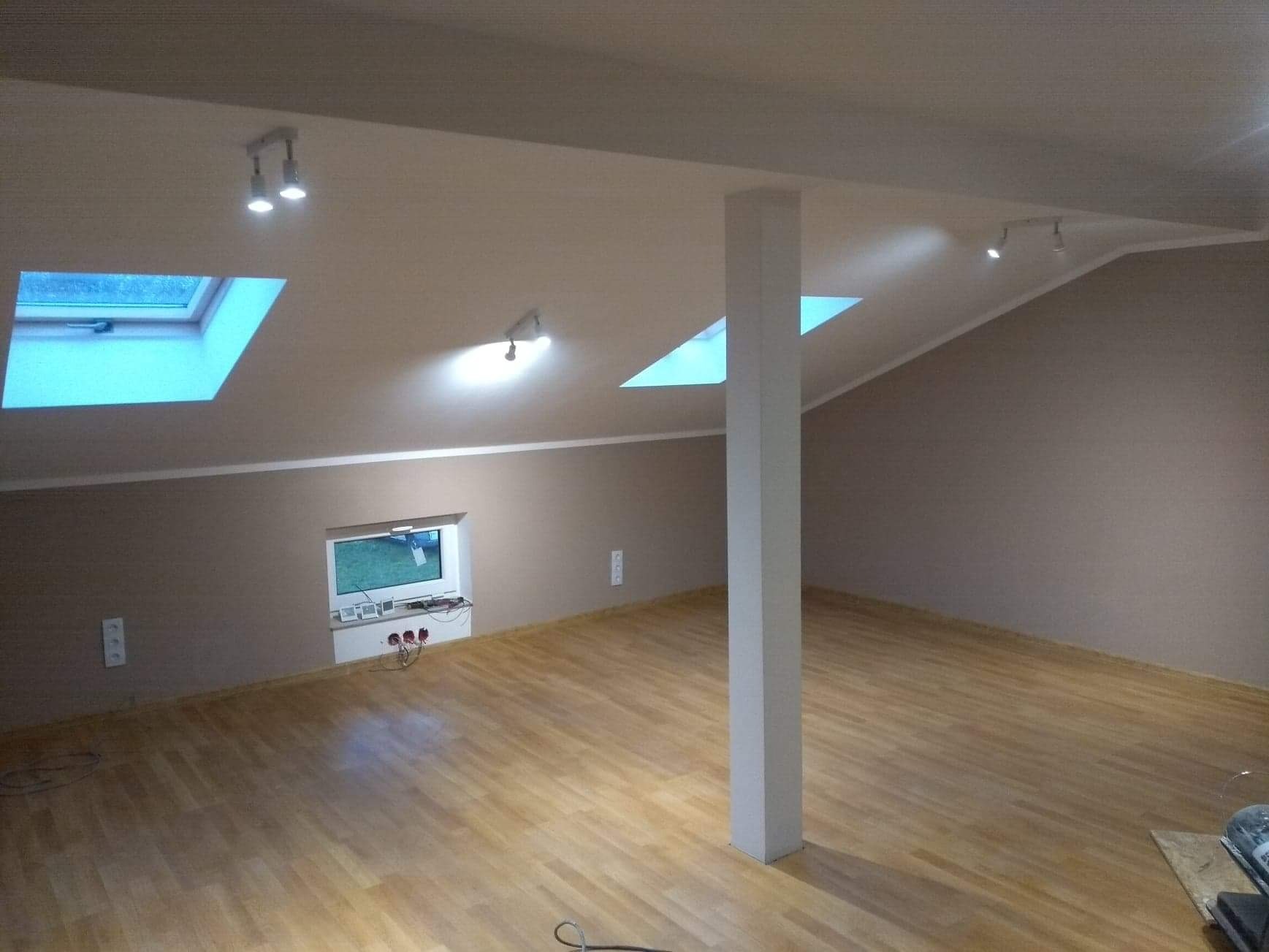 Poddasze z oknami dachowymi i oświetleniem punktowym, świeżo pomalowane ściany w odcieniach beżu i jasna drewniana podłoga, widoczny słup nośny.