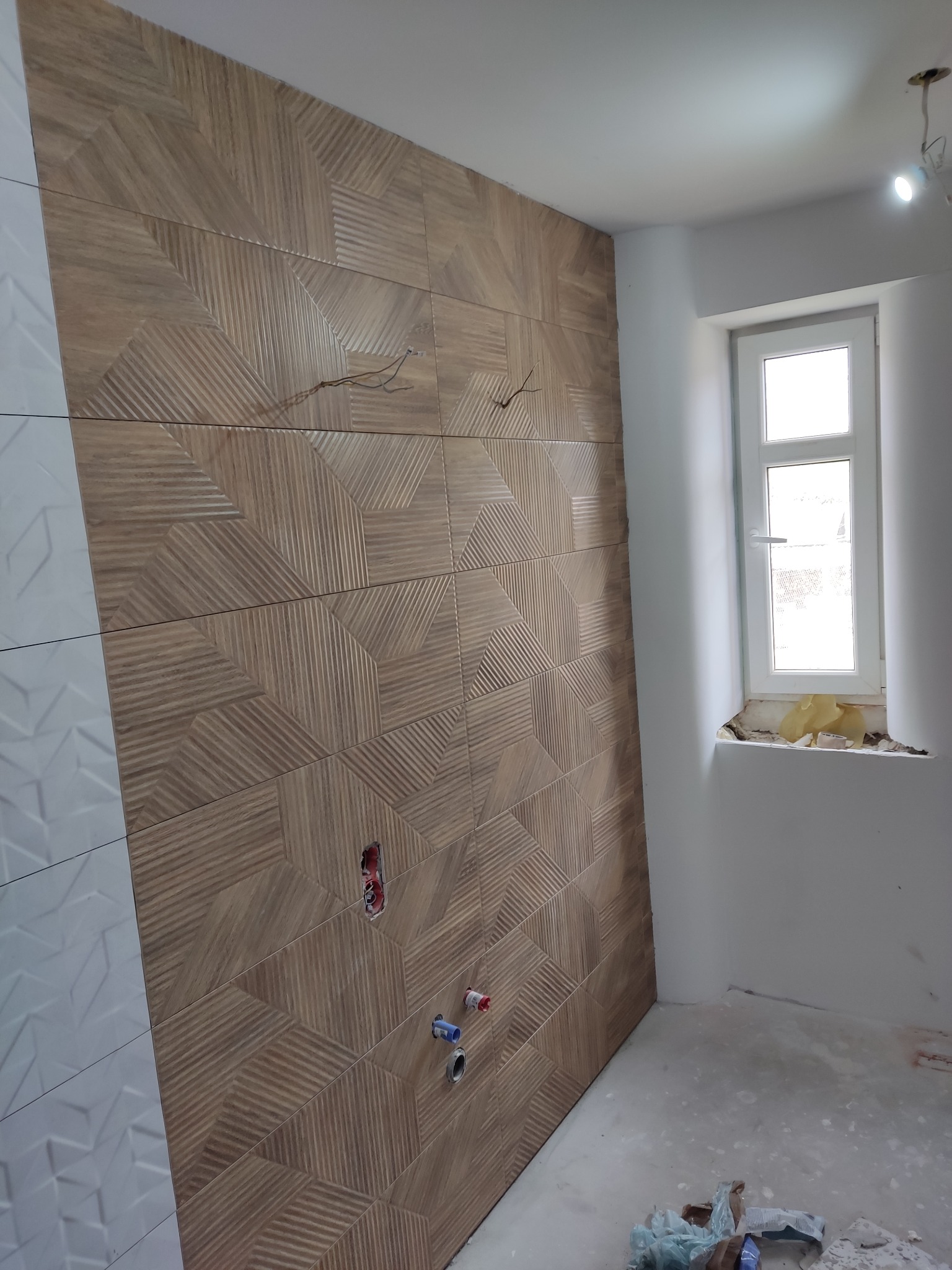 Ściana w trakcie wykańczania z dekoracyjnymi, drewnopodobnymi płytkami oraz białą, wzorzystą glazurą, widoczne wystające kable i rury, surowy stan podłogi i okna.