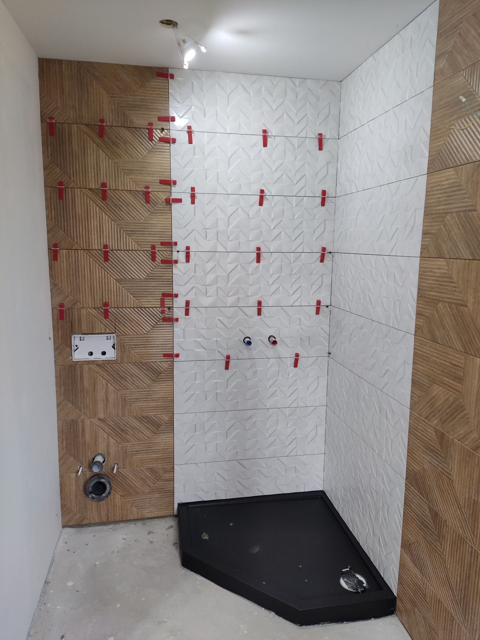 Łazienka w trakcie wykańczania: montaż płytek drewnopodobnych i białych z geometrycznym wzorem, widoczne krzyżyki poziomujące, czarny brodzik i instalacja podtynkowa WC.