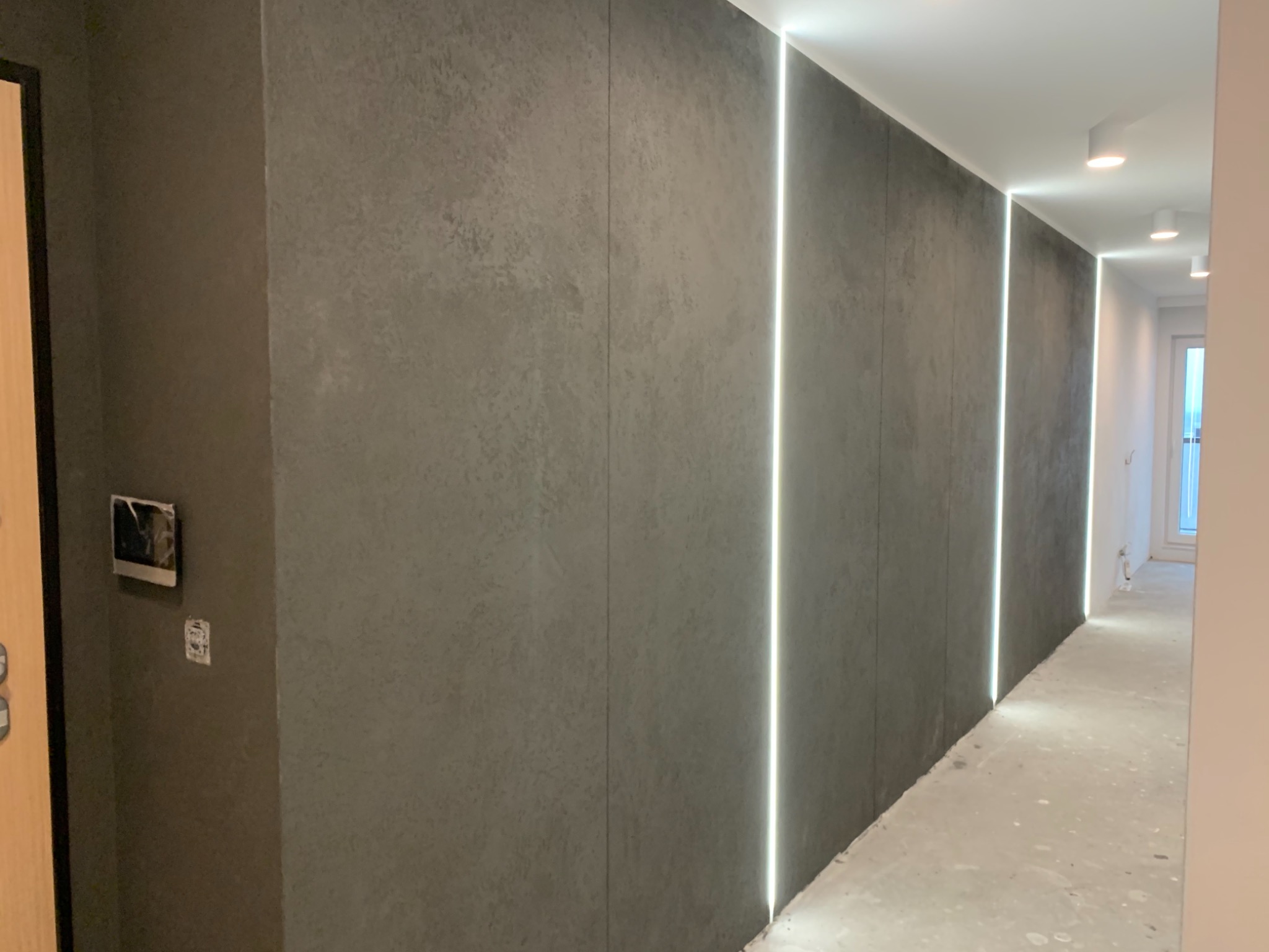 Korytarz z szarymi panelami ściennymi o betonowej fakturze, podświetlonymi pionowymi pasami LED, widoczne lampy sufitowe i fragment drzwi.