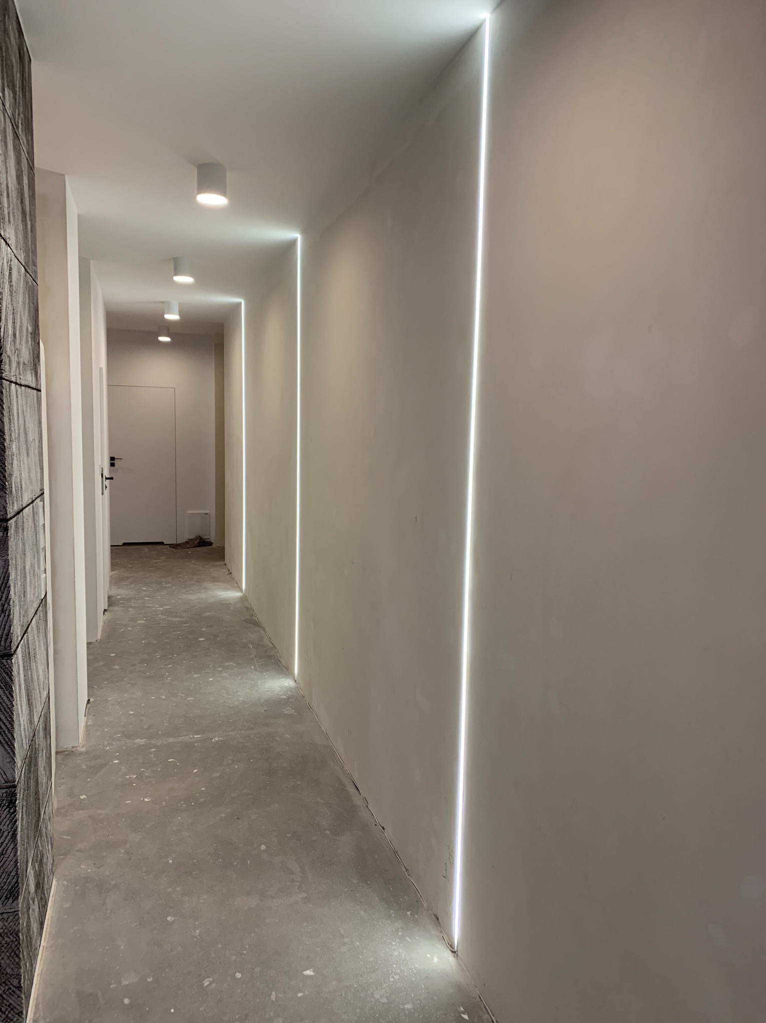 Korytarz w trakcie prac wykończeniowych z pionowymi wnękami oświetleniowymi w ścianach i sufitowymi lampami punktowymi.