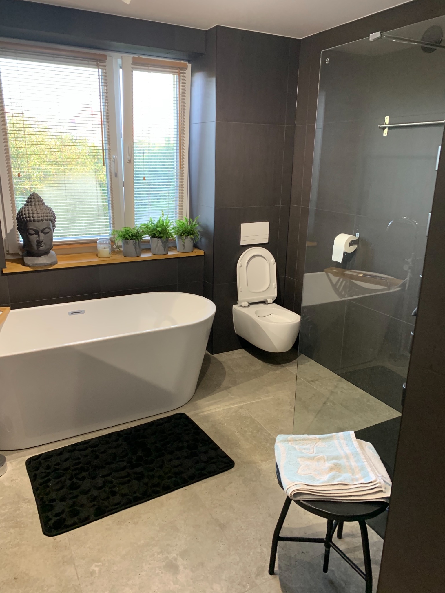 Nowoczesna łazienka z wolnostojącą wanną, podwieszaną toaletą, kabiną prysznicową ze szklaną ścianką, szarą glazurą i drewnianym blatem z roślinami doniczkowymi pod oknem.