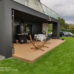 Zadaszony taras z desek w kolorze jasnego drewna, wyposażony w grill, białą sofę i hamak na tle zadbanego trawnika i nowoczesnego domu.