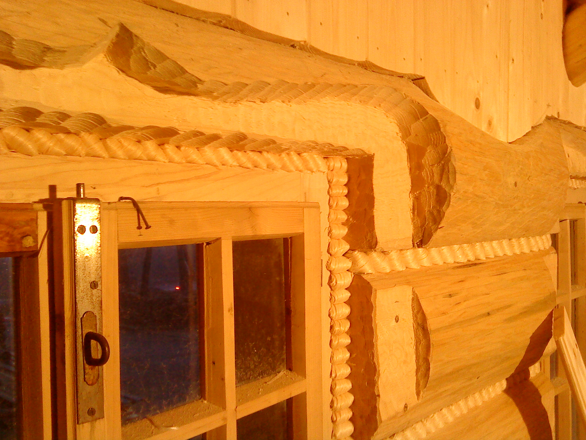 Fragment ściany domu z bali, z widocznym narożnikiem i oknem z metalowym zamknięciem, dekoracyjnie obrobione drewno z snycerskimi zdobieniami.