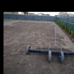 Przygotowany teren pod trawnik z użyciem walca do wyrównywania, widoczny rząd małych tuj wzdłuż ogrodzenia.