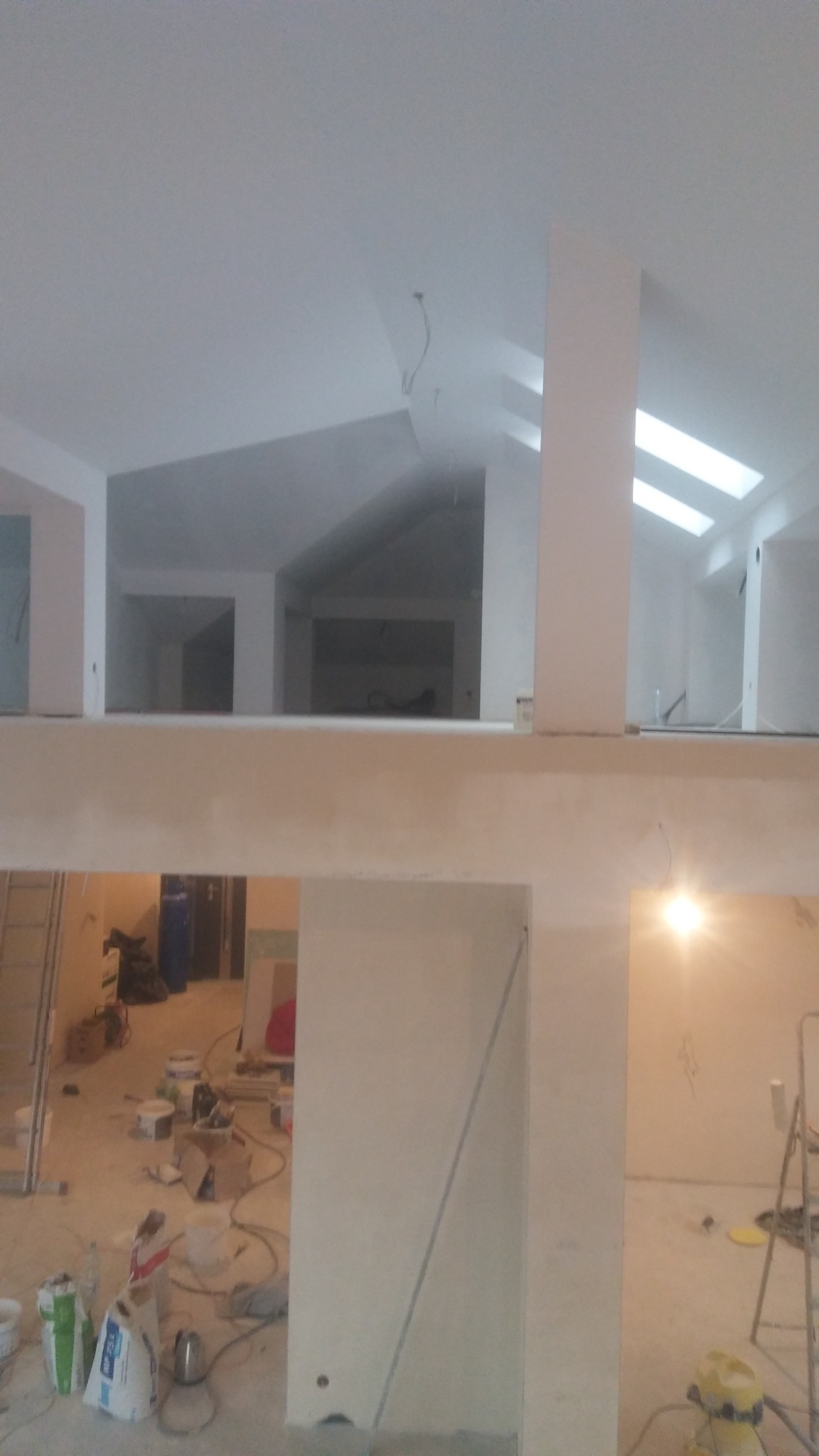 Wnętrze dwupoziomowego domu w trakcie prac wykończeniowych; widoczne gładkie ściany, sufit z oknami dachowymi oraz materiały budowlane na podłodze.