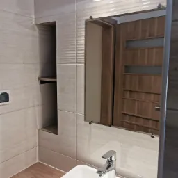 Nowoczesna łazienka z białymi płytkami imitującymi kamień, lustro z oświetleniem, umywalka nablatowa i drewnopodobna podłoga; w lustrze odbicie drzwi z szybą.