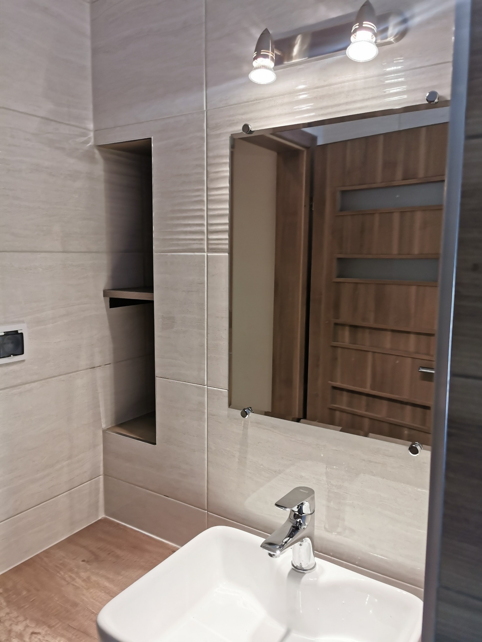 Nowoczesna łazienka z białymi płytkami imitującymi kamień, lustro z oświetleniem, umywalka nablatowa i drewnopodobna podłoga; w lustrze odbicie drzwi z szybą.