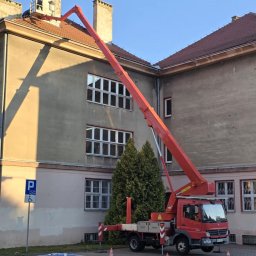 Usługi Podnośnikami Koszowymi Krzysztof Miłoński. - Czerwony pojazd z wysięgnikiem parkuje w Komorowie, przy budynku z ceglanym dachem i kominami, gdzie na platformie wysięgnika znajduje się osoba pracująca przy kominie.